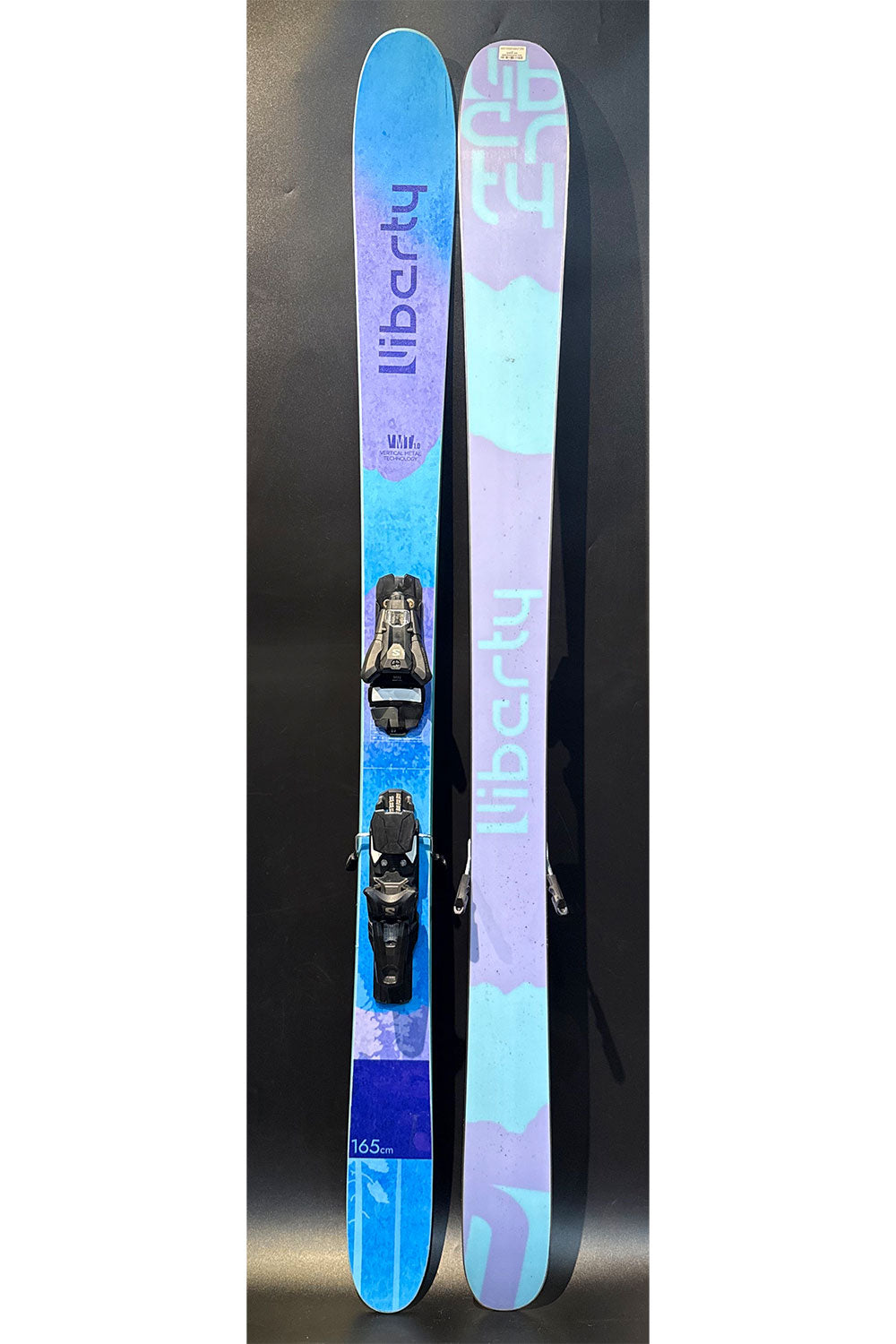 Pair of blue and purple Liberty skis with bindings on a dark background