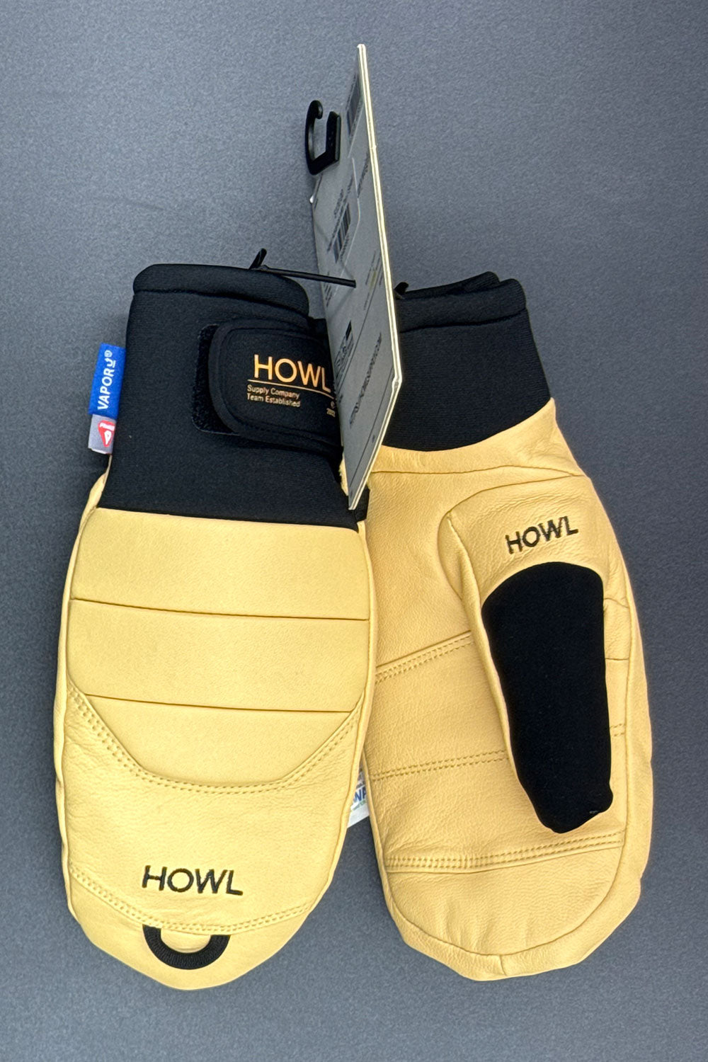 Pair of tan mittens with black accents on a gray background, featuring the brand 'HOWL'.
