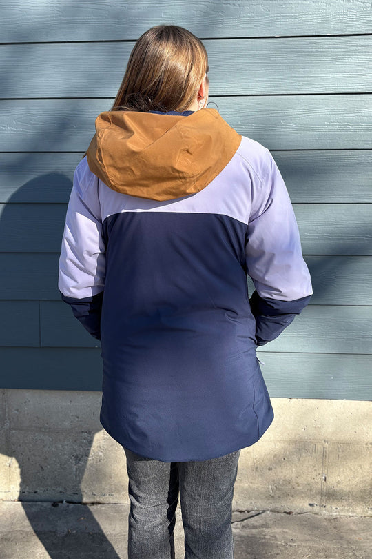 Person wearing a colorful jacket with a brown hood against a gray wall.