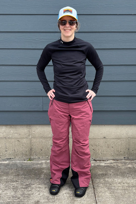 Person wearing black long-sleeve shirt, maroon pants, and a colorful cap standing against a blue wall.
