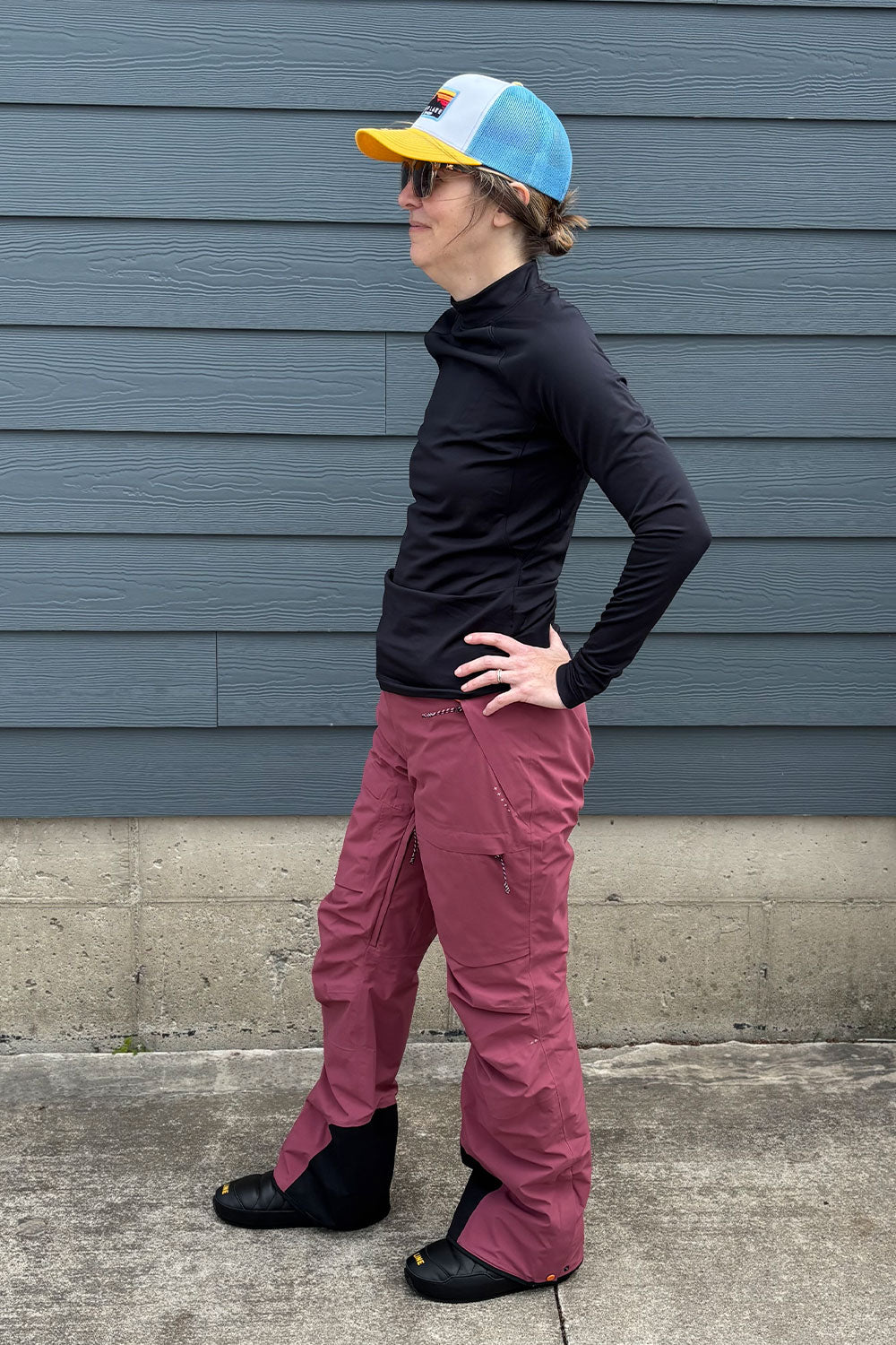 Person wearing a black long-sleeve shirt, pink pants, and a colorful cap against a blue wall.