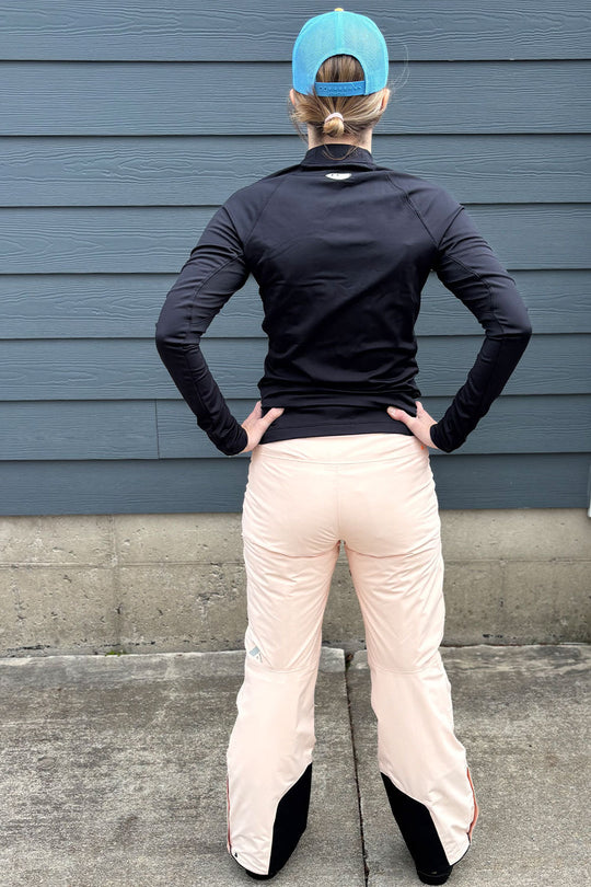 Person wearing a black jacket and light pink pants with a blue cap, standing against a gray wall.