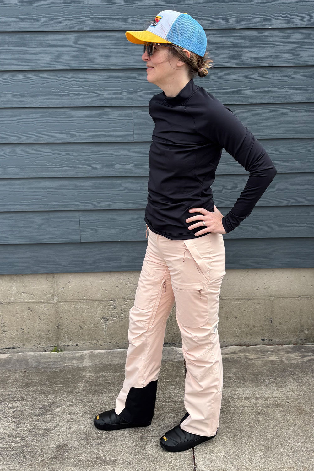 Person wearing a black turtleneck, light pink pants, and a colorful cap against a blue wall.