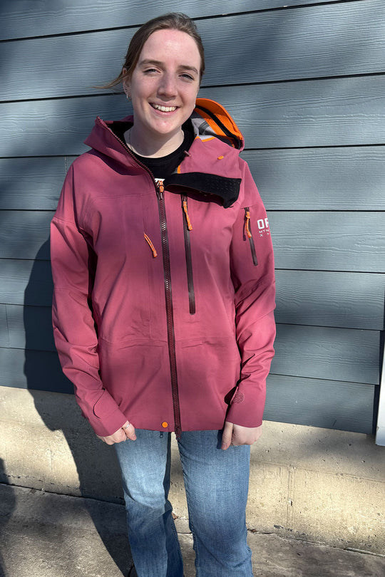 Person wearing a pink jacket with a brand logo, standing against a gray wall.