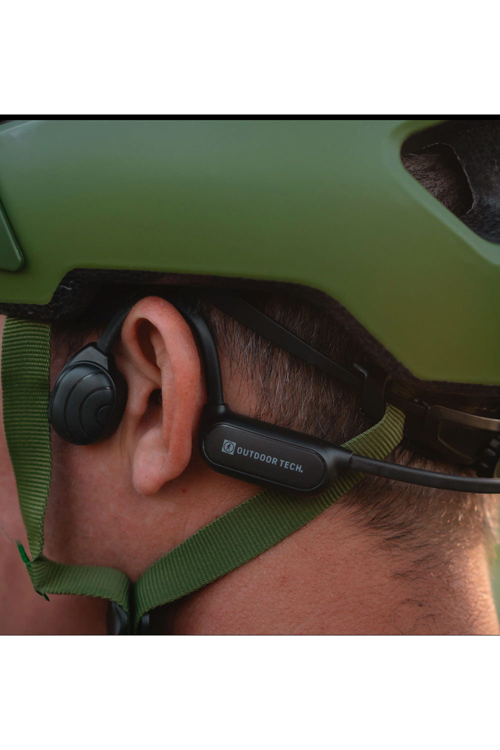 Person wearing a green helmet with a black device on their ear, branded 'Outdoor Tech'.