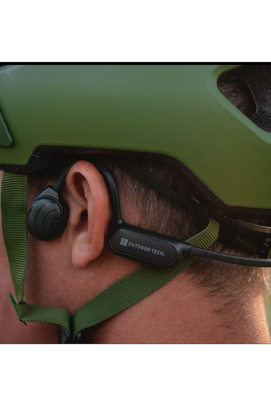 Person wearing a green helmet with a black device on their ear, branded 'Outdoor Tech'.