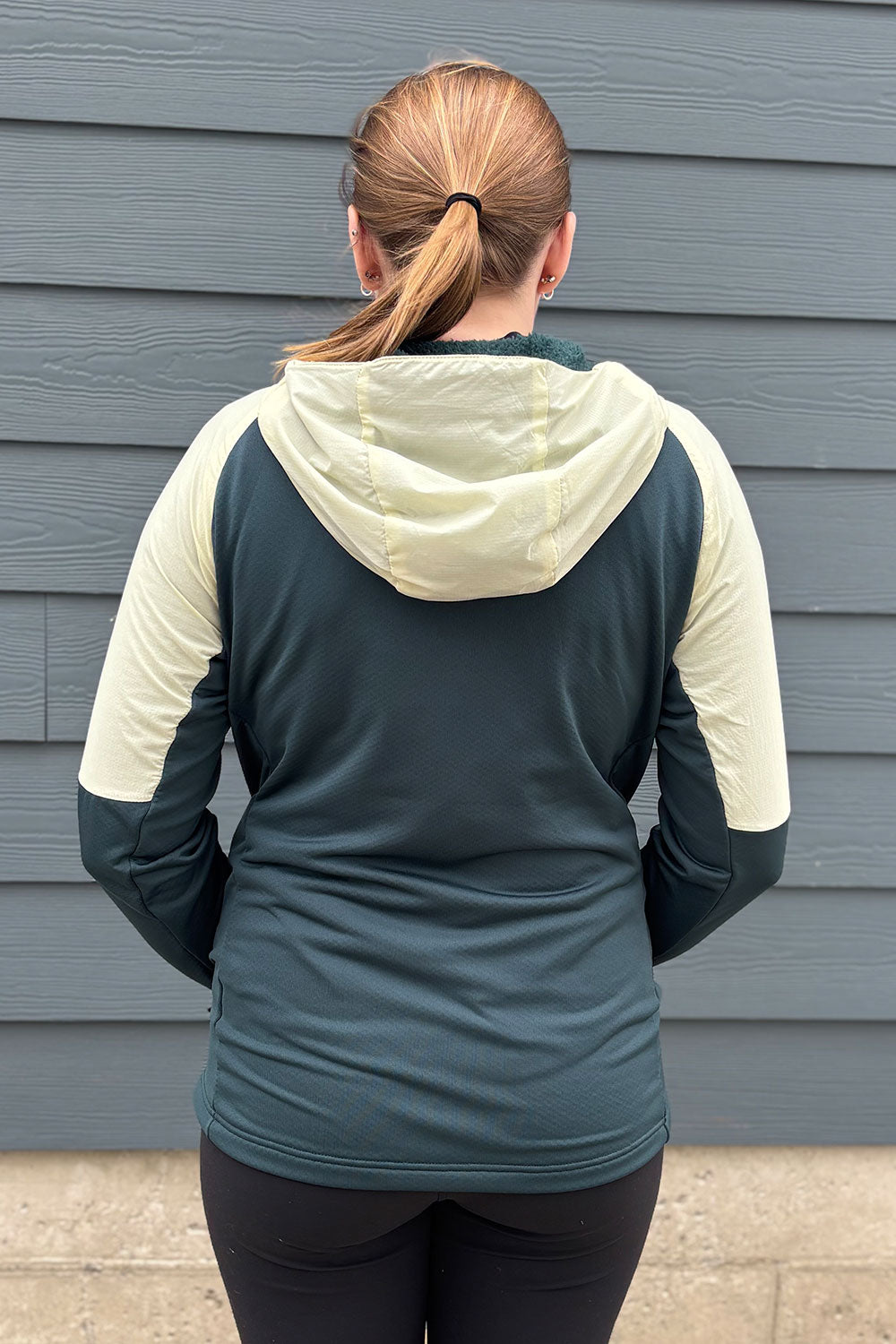 Person wearing a teal and beige hoodie standing against a gray wall.