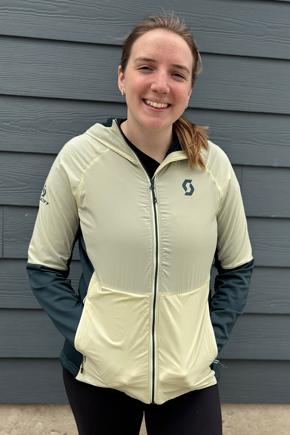 Person wearing a light-colored jacket with a brand logo against a gray wall.