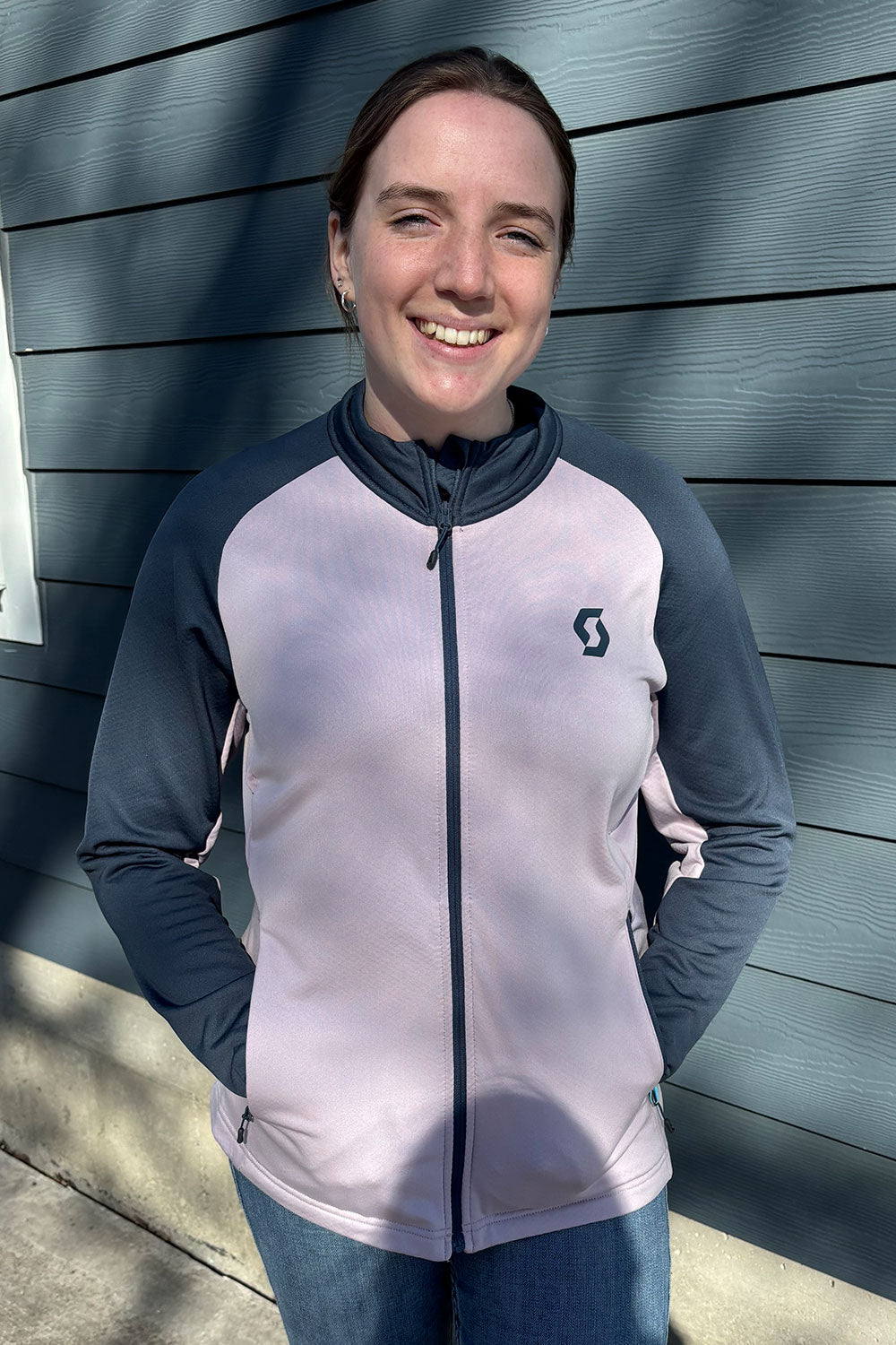Person wearing a gray and white jacket with a logo, standing against a blue wall.