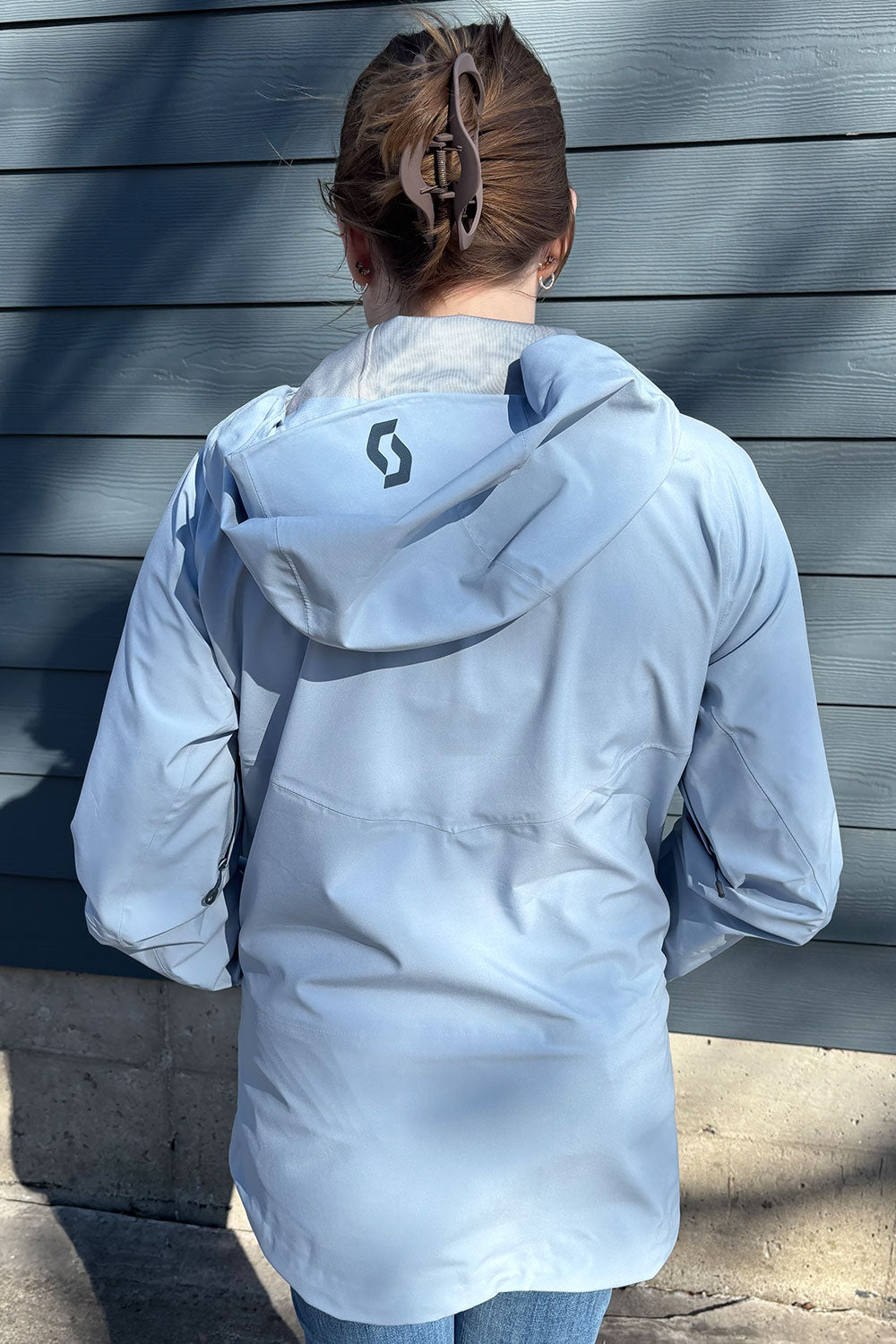 Person wearing a light blue jacket with a brand logo, standing against a gray wooden wall.