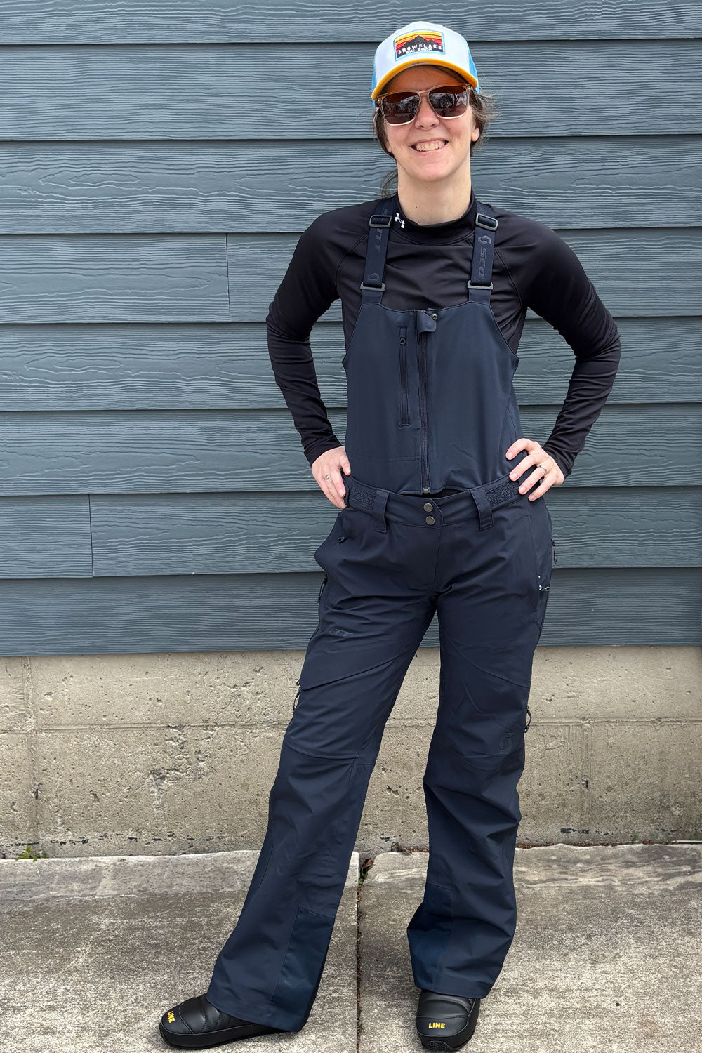 Person wearing navy overalls and a black long-sleeve shirt standing against a gray wall.