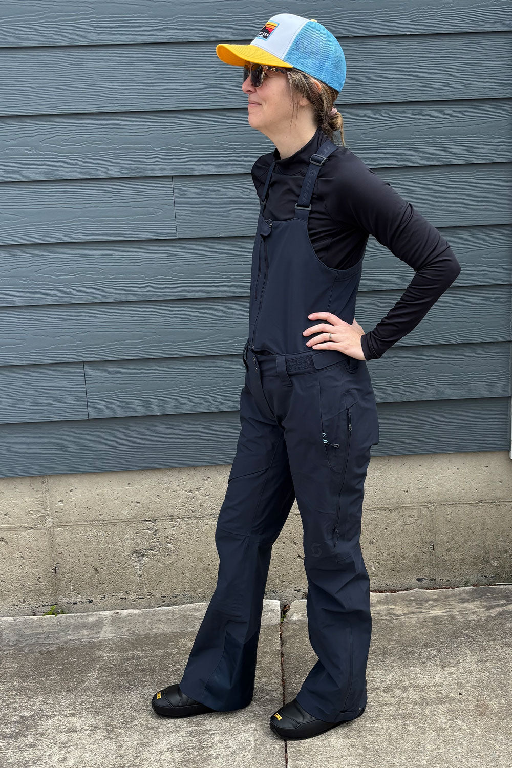 Person wearing a black jumpsuit and colorful cap standing against a blue wall.