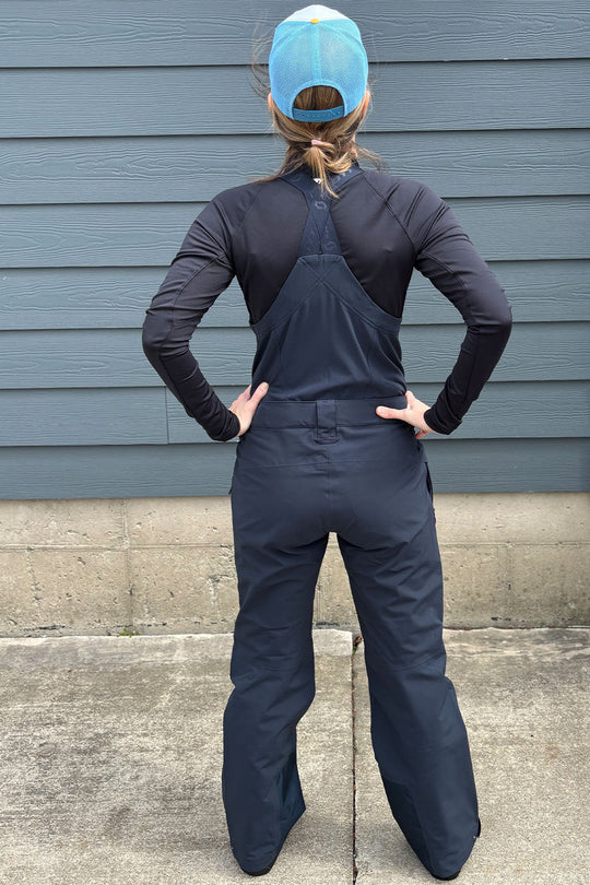 Person wearing a black long-sleeve shirt and blue overalls standing against a gray wall.