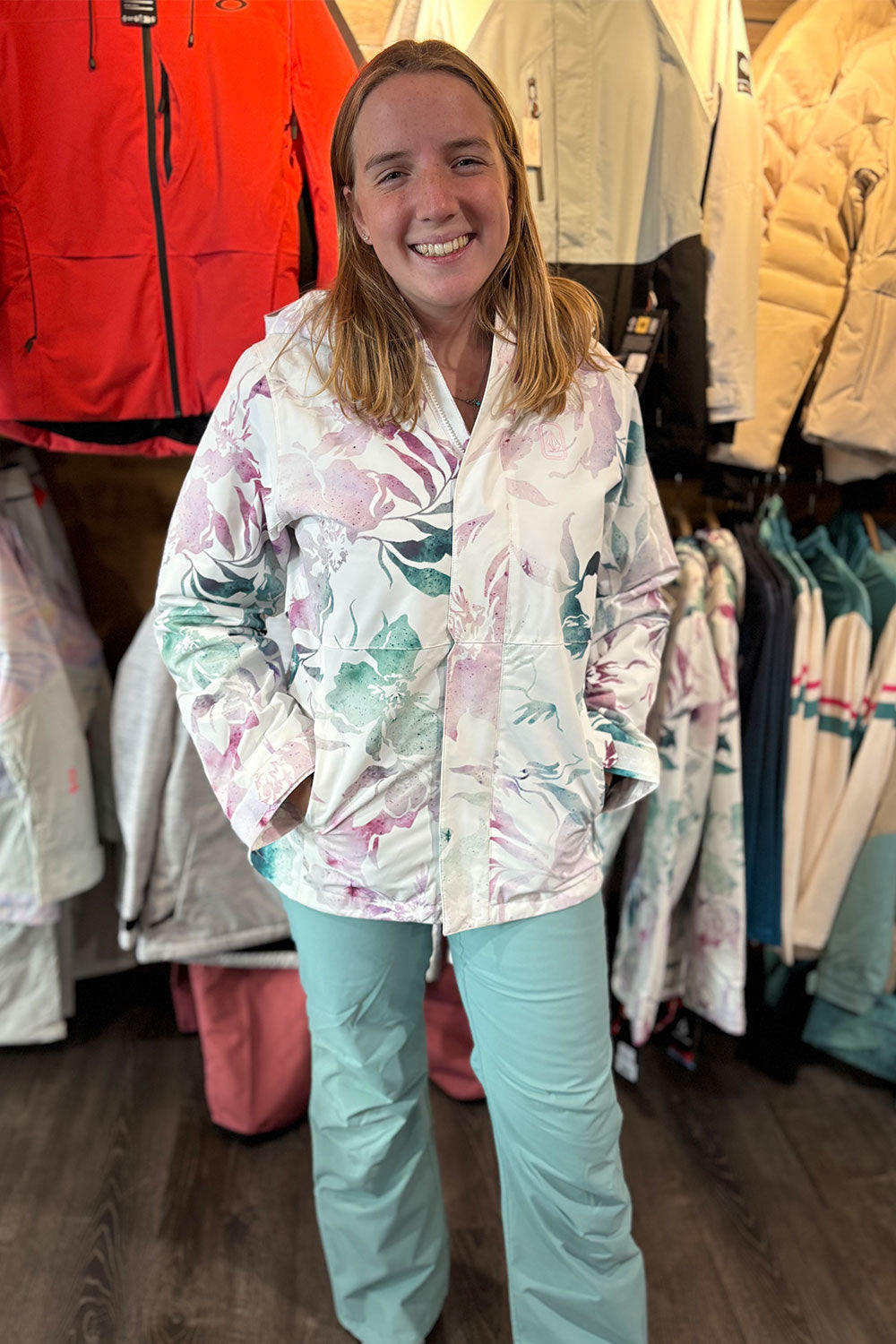 Woman wearing a colorful jacket and light blue pants standing in a clothing store with racks of jackets behind her.