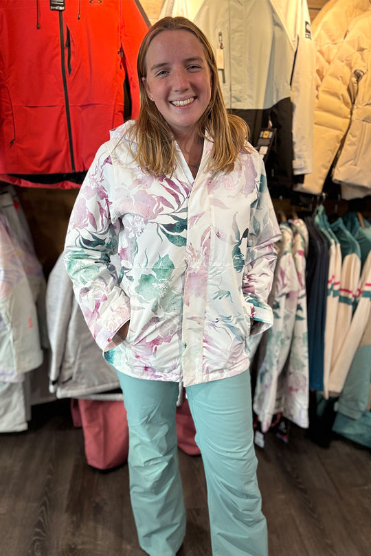 Woman wearing a colorful jacket and light blue pants standing in a clothing store with racks of jackets behind her.